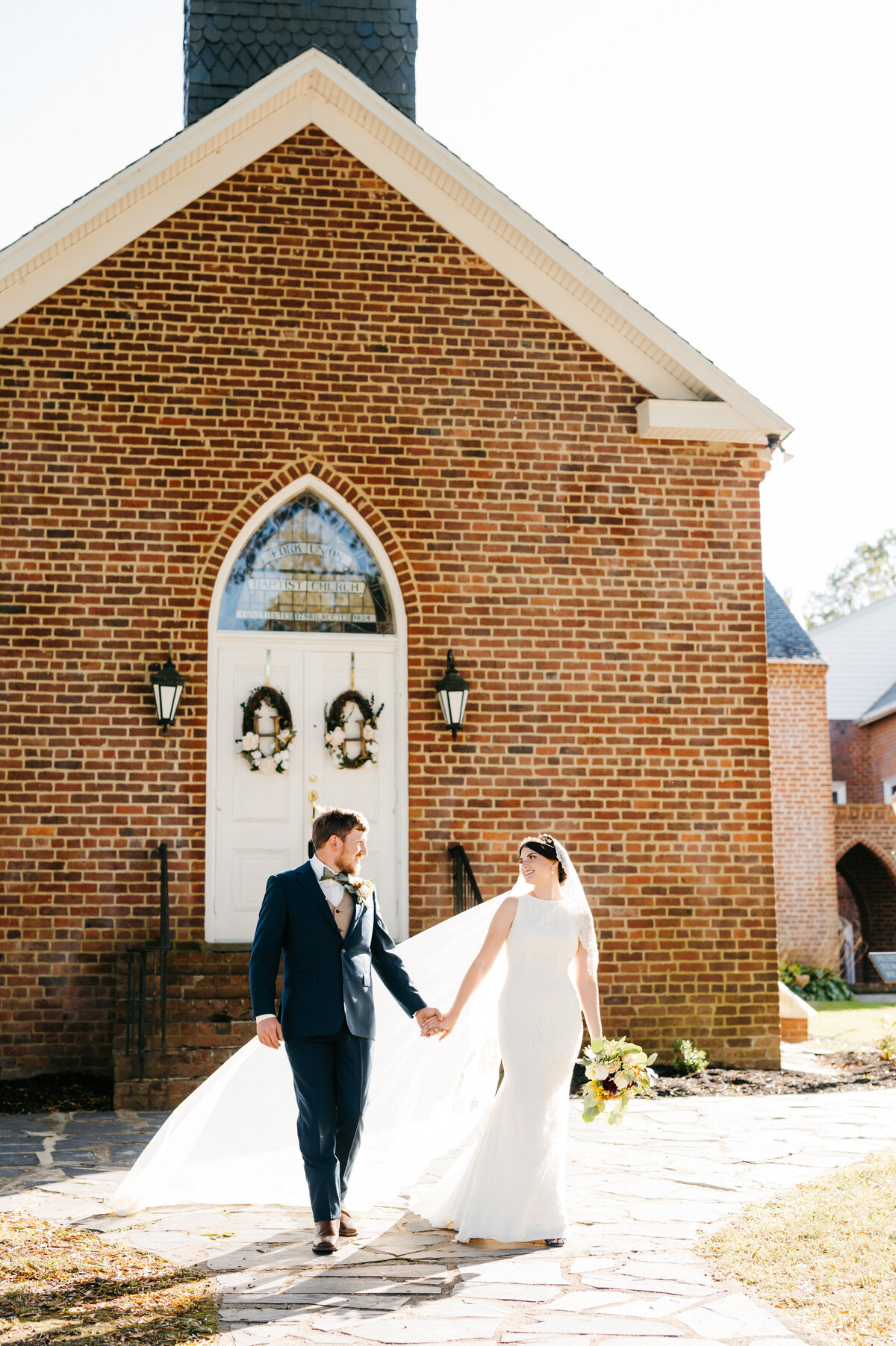 Richmond Wedding venues with brick chapel and stone paths featuring bride and groom walking together while holding hands at sunset with the brides veil illuminating as the sun shines through it while it blows in the wind