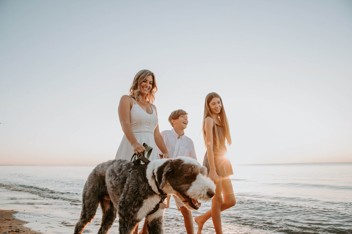 Family photoshoot at Grand Bend beach - Mom and tween son and daughter are walking their sheepdog in the shoreline at sunset. Mom and children are laughing.