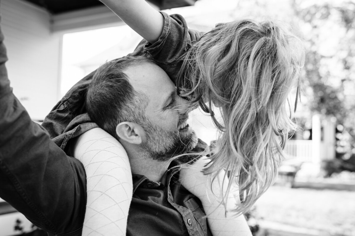 A black and white image of a father and daughter sharing a playful moment outside. The daughter is sitting on the father’s shoulders, and they are both smiling with their faces close together.