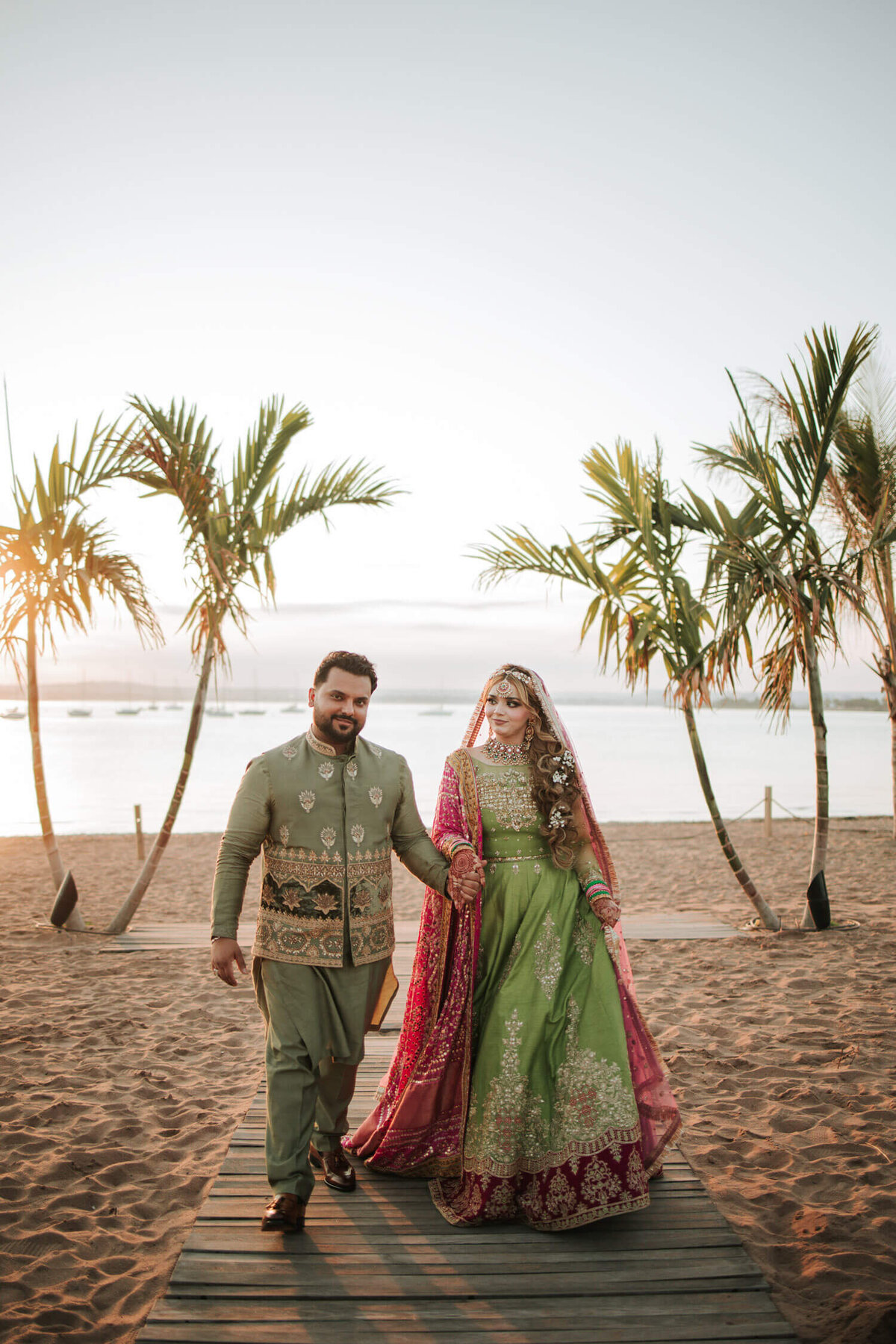 A couple in vibrant traditional attire walks hand-in-hand on a sunlit beach, surrounded by palm trees and golden light. At Antony's Pier Anthony's Ocean View New Haven, CT