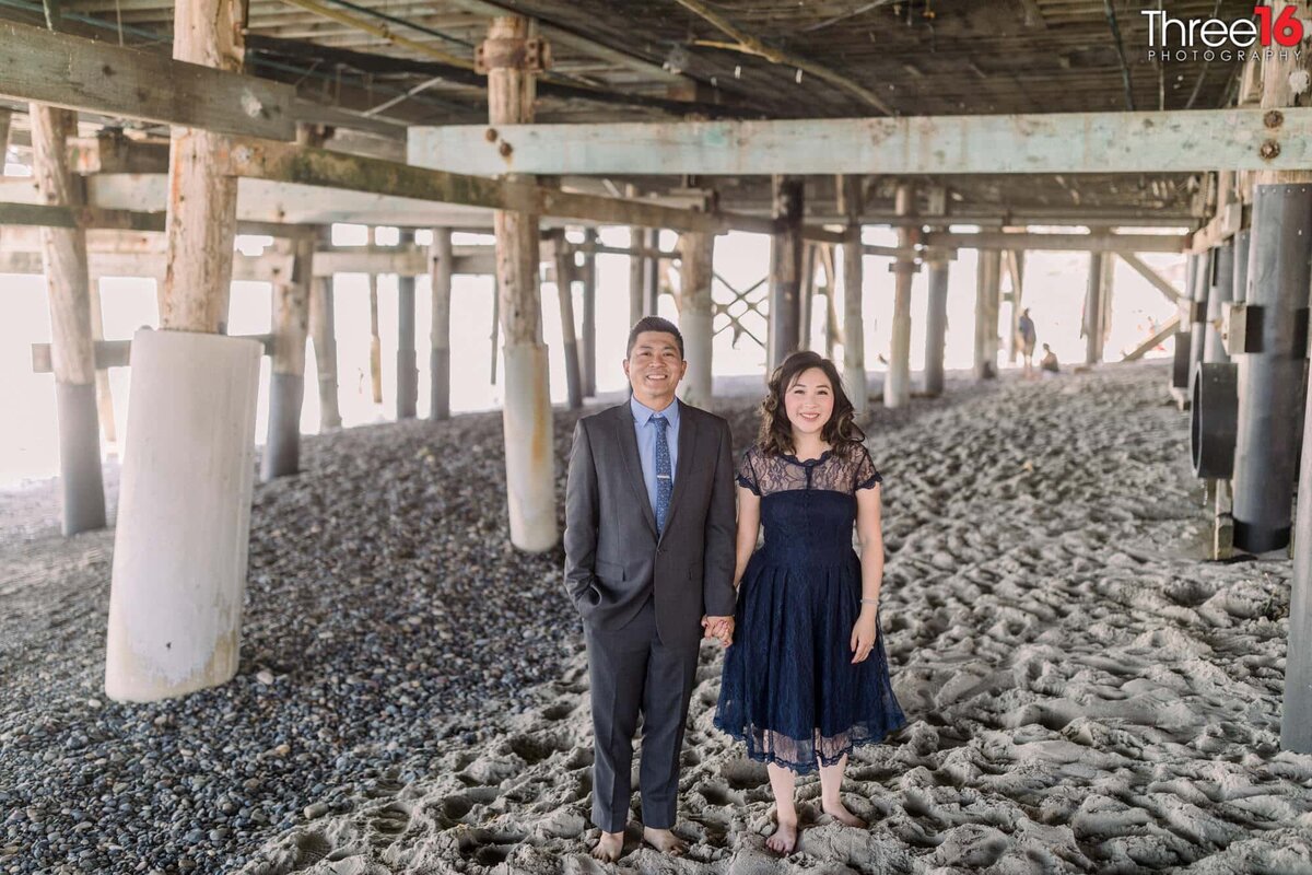 Engaged couple hold hands as they stand under the San Clemente Pier for engagement photos