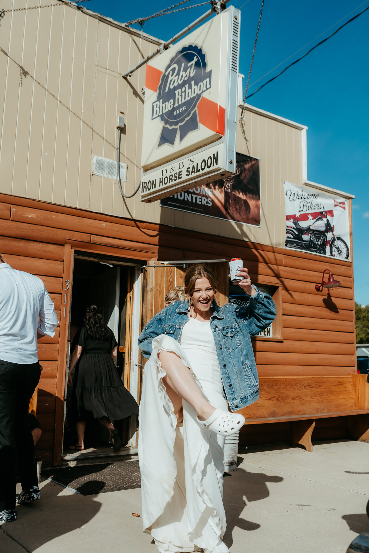 Bride in denim jacket and Crocs celebrating outside small-town Minnesota bar wedding venue.