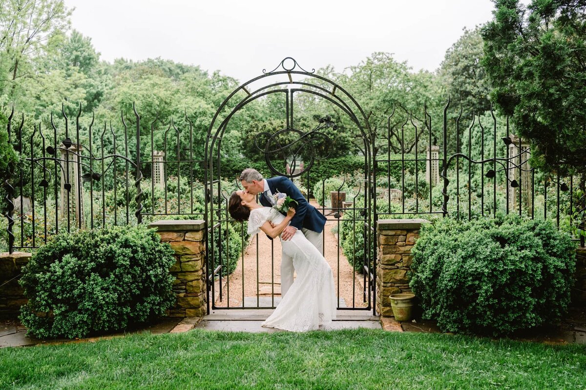 Groom kisses and dips bride in front of gate at gardens at Waterperry Farm wedding
