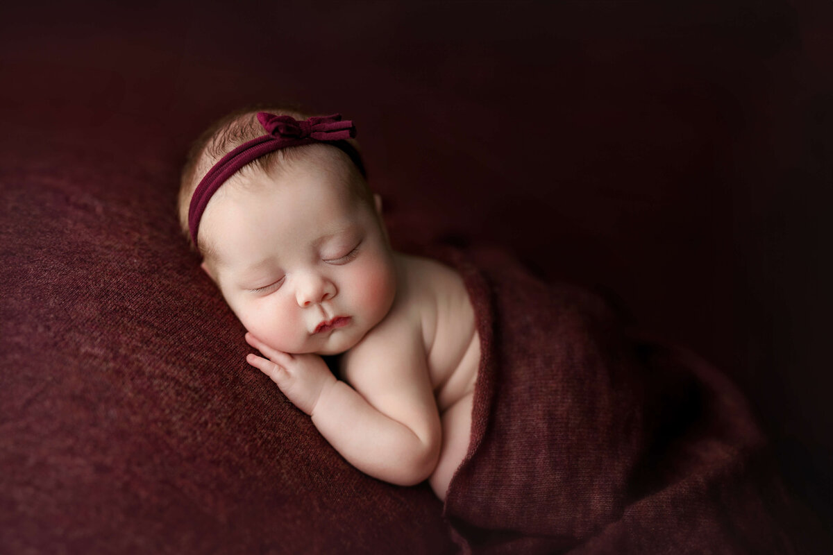 newborn baby girl laying on her tummy  on a maroon backdrop with matching headband and wrap at a newborn photo shoot in Northern VA