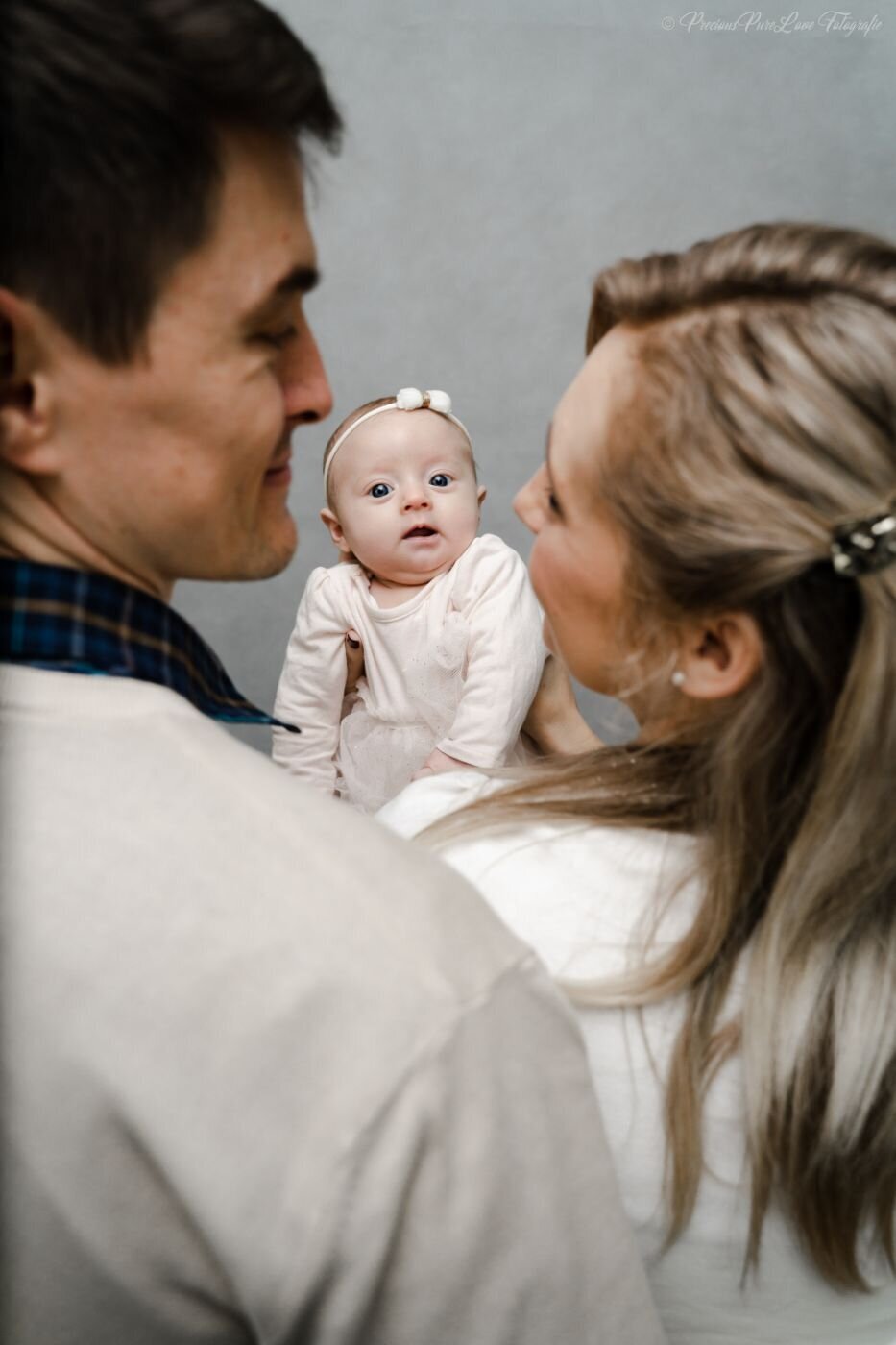  A black and white, eye-level photo of a newborn baby being held up between two parents. The parents are out of focus in the foreground, looking at the baby. The baby is in sharp focus, looking directly at the camera with wide eyes. The baby is wearing a white outfit and a small bow on its head.