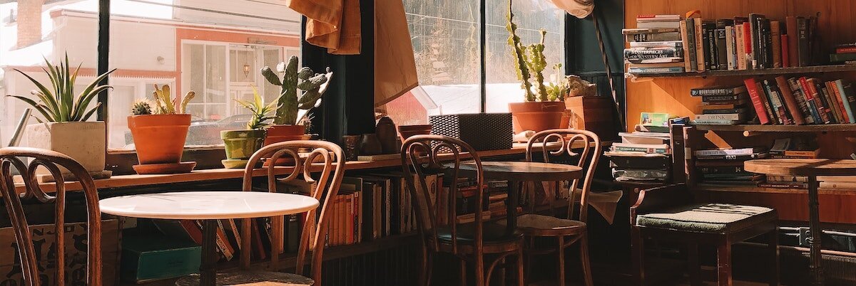 Warm-toned cafe with two-top tables in front of a large window and stacks of books on shelves. Photo by Haley Black via Pexels.