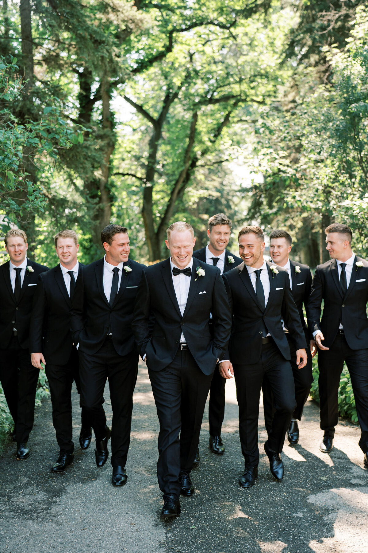 A group of men in black suits and ties walk together on a tree-lined path, perhaps discussing full service wedding planner options.