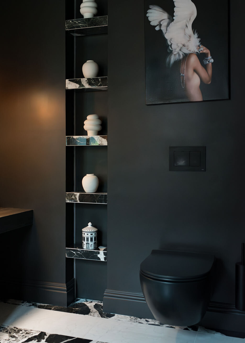 A modern bathroom with dark walls and a black toilet. There are built-in mirrored shelves next to the toilet, holding decorative items including white vases and a small white sculpture. A dark framed art piece featuring a person and white bird is on the wall.