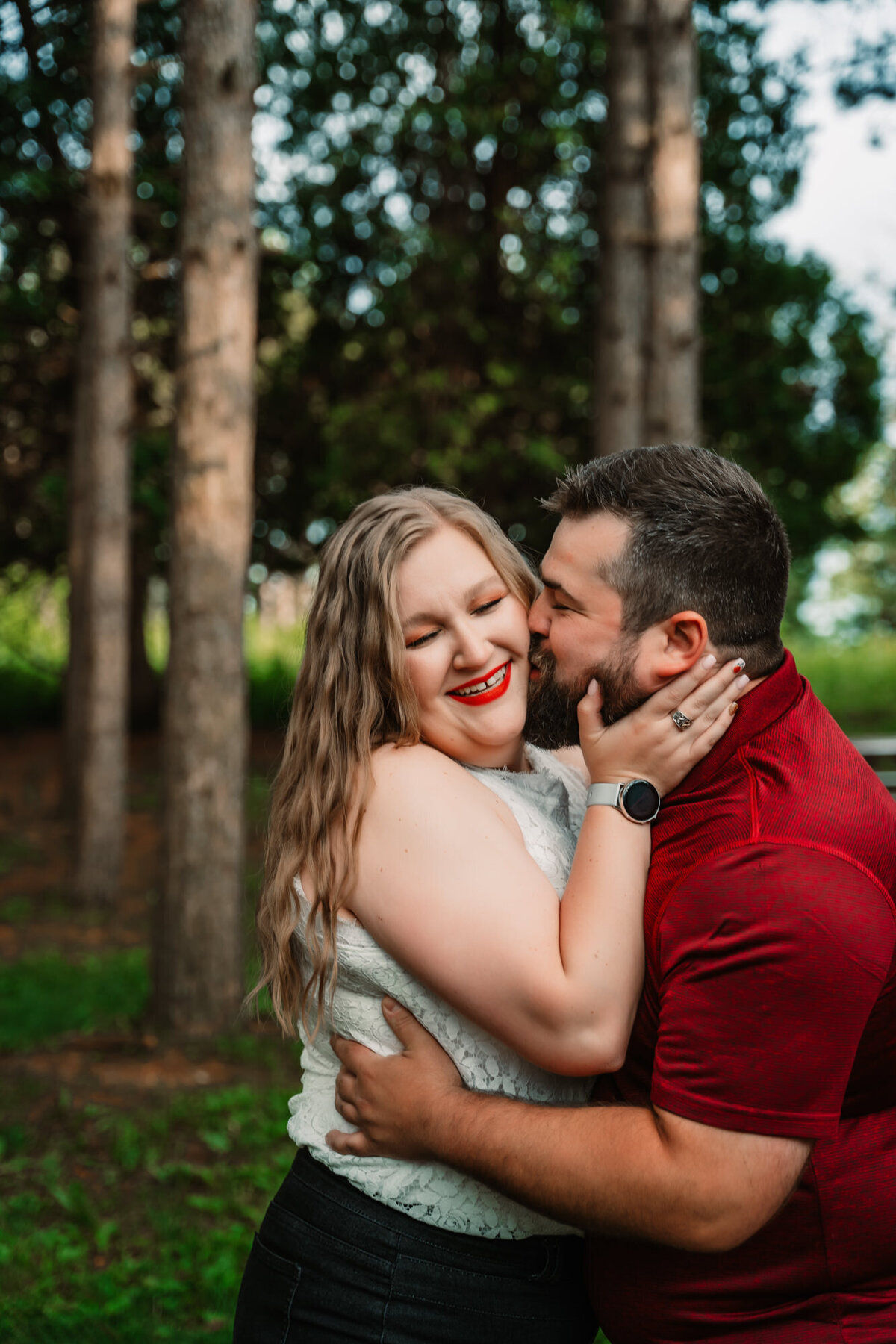 man kisses woman on cheek while she smiles in a forest