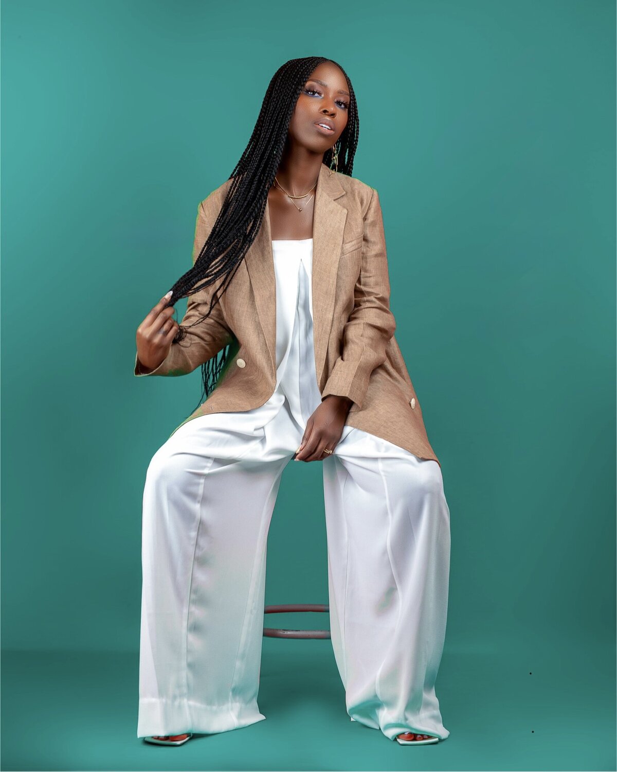 H.B Lighthouse founder Genevia Sawyer poses with Fulani braids wearing a brown linen blazer and white chiffon jumpsuit, sitting on a stool