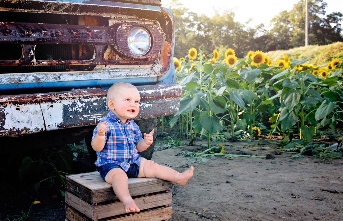 baby boy in plaid shirt sitting on wooden crate in front of an old rusty pickup truck in a sunflower field