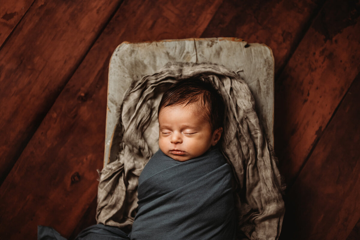 newborn photos in Maryland with baby in a dark grey swaddle on a miniature bed  on a red oak wood floor