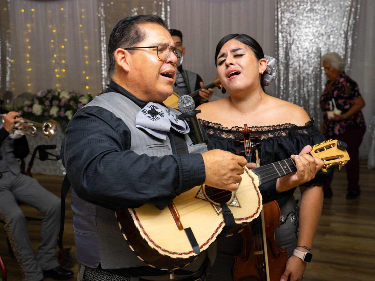 Two mariachi singers, male and female, gather around a guitar in a sparkling, shadowy ballroom and sing together with eyes clothes mouths open in an emotional refrain. Photo by SAVI Photography - Photographer in Riverside