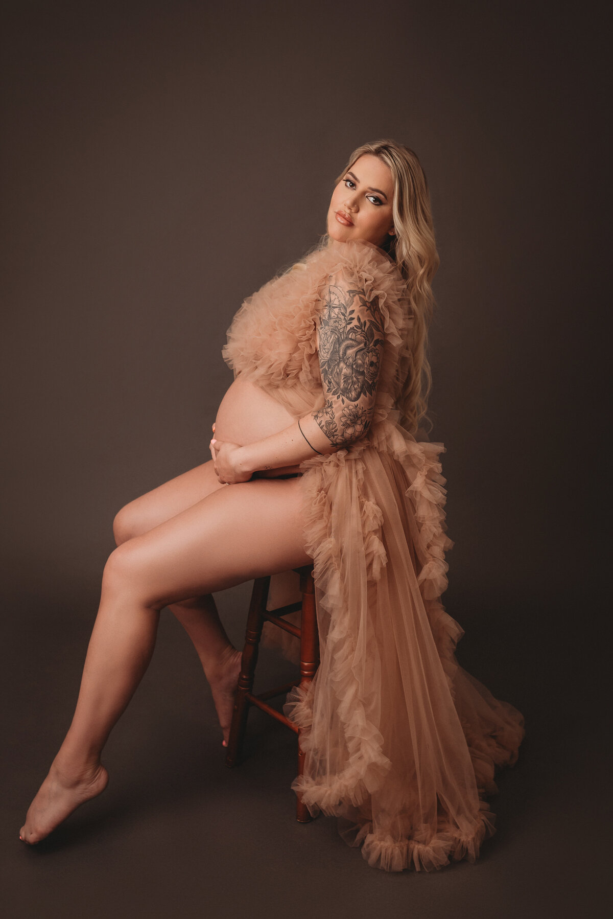 Atlanta, Georgia newborn photographer portrait session with pregnant woman at maternity session sitting on stool on dark grapy backdrop holding baby bump wearing tulle ruffle maternity dress