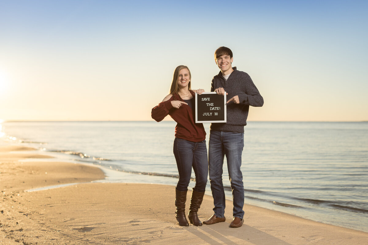 Engagement photos for a couple taken at Ft. Morgan, Alabama at sunset on the beach holding a save the date board.