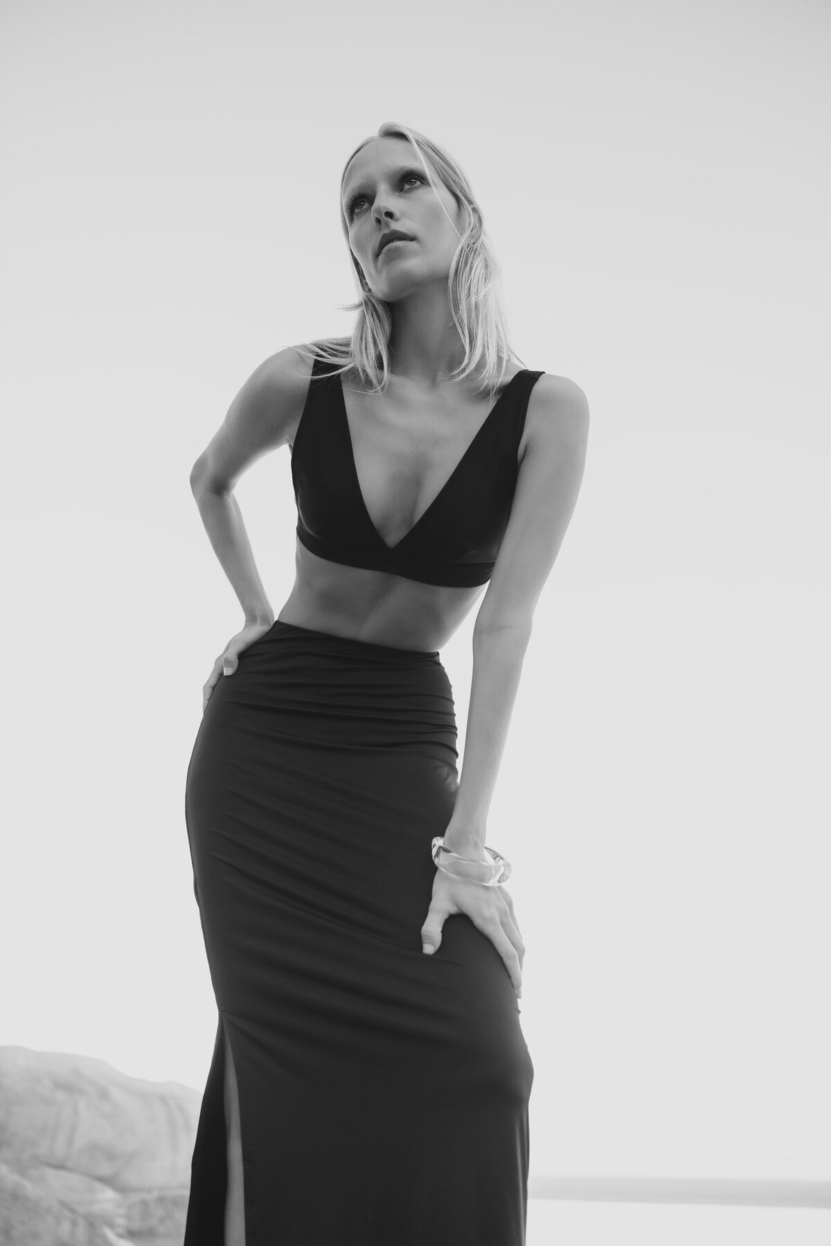 Zwart-wit portret van model Johanna in een elegante cut-out jurk, poserend met krachtige houding en opwaartse blik, gefotografeerd tegen een minimalistische lucht en ruige kustlijn in Kaapstad.