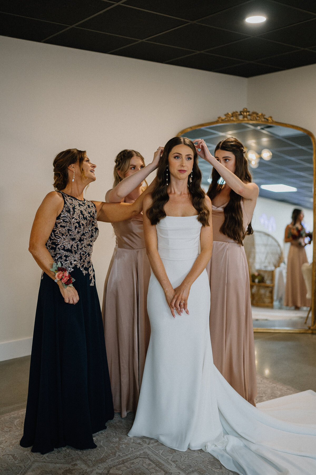 Photo of Two sisters and mother of the bride helping to place the veil on an elegant bride in a slimming white dress and pearl earrings by Destiny Rae Photography