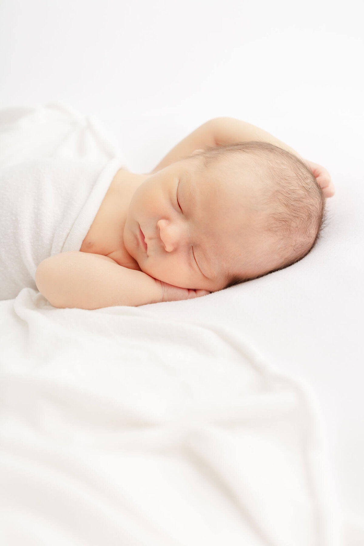 Newborn baby sleeping tucked under a white sheet with hands up by head.