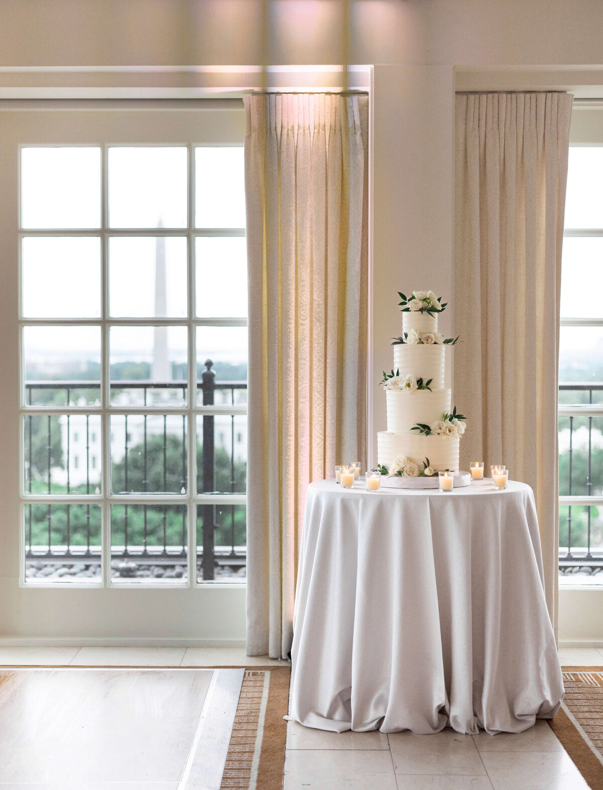 A three-tiered white wedding cake adorned with flowers sits on a round table draped in white cloth. The table is surrounded by small candles. Large windows in the background reveal a blurred cityscape view.