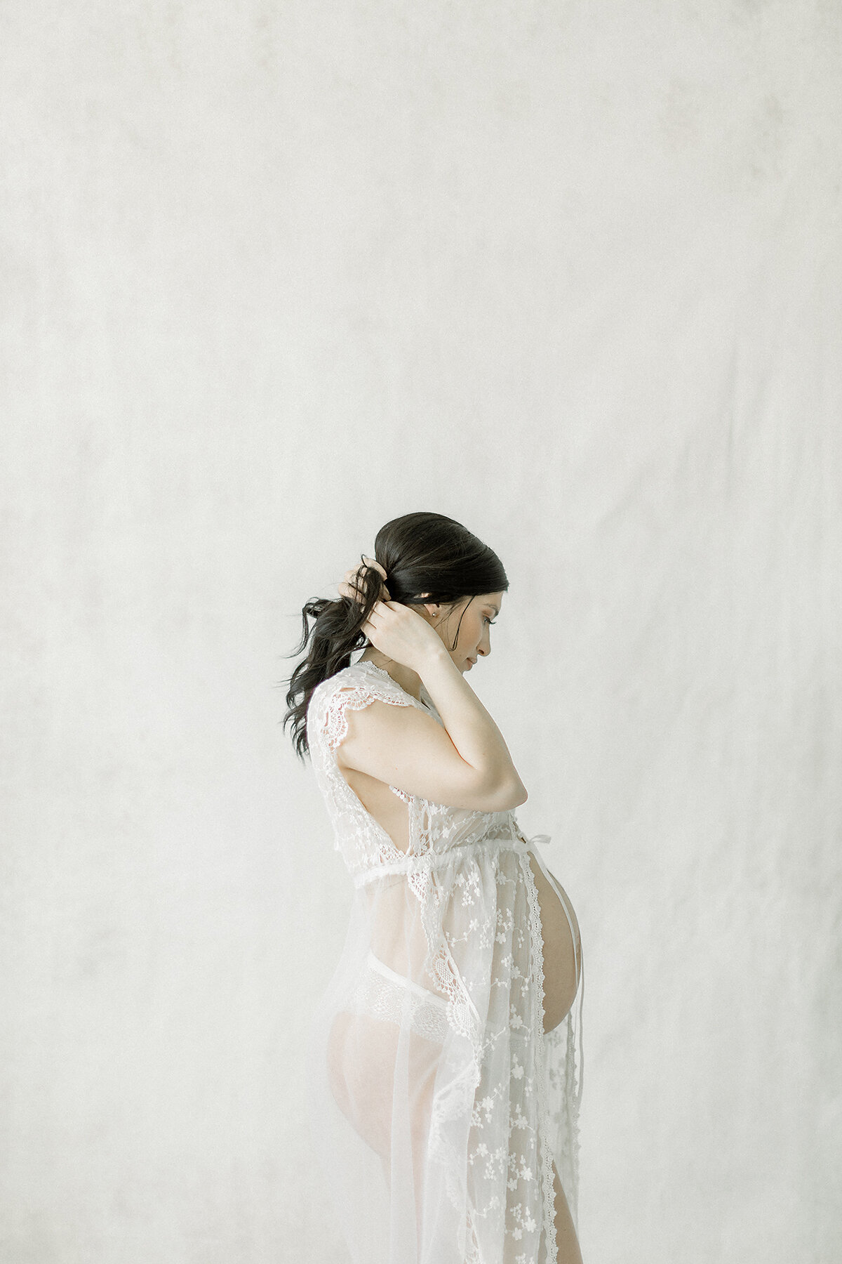 A light filled photo of an in studio maternity session of a pregnant DFW mother standing by the wall as she is elegantly holding her hair up and away from her face.