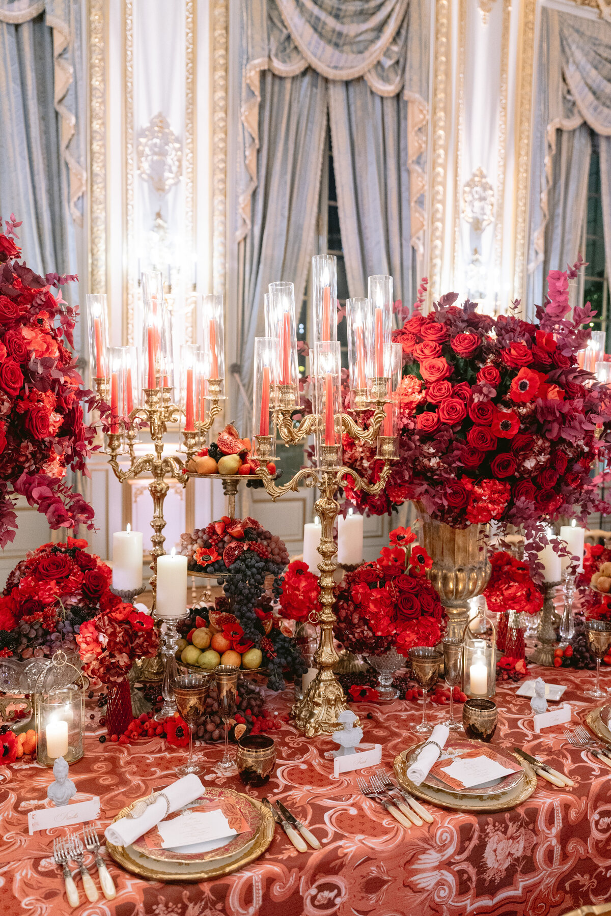 The stunning photo was taken at the luxurious Shangri La Hotel & Ritz Paris Hotel in Paris, France, capturing a lavishly decorated dining table by photographer Matteo Coltro and Zen Film Works as Videographers. The table features ornate gold candle holders adorned with tall red candles, abundant arrangements of exquisite red roses, and intricate place settings with elegant gold-rimmed plates. The backdrop showcases opulent drapes with intricate details, further enhancing the grand ambiance of the scene.