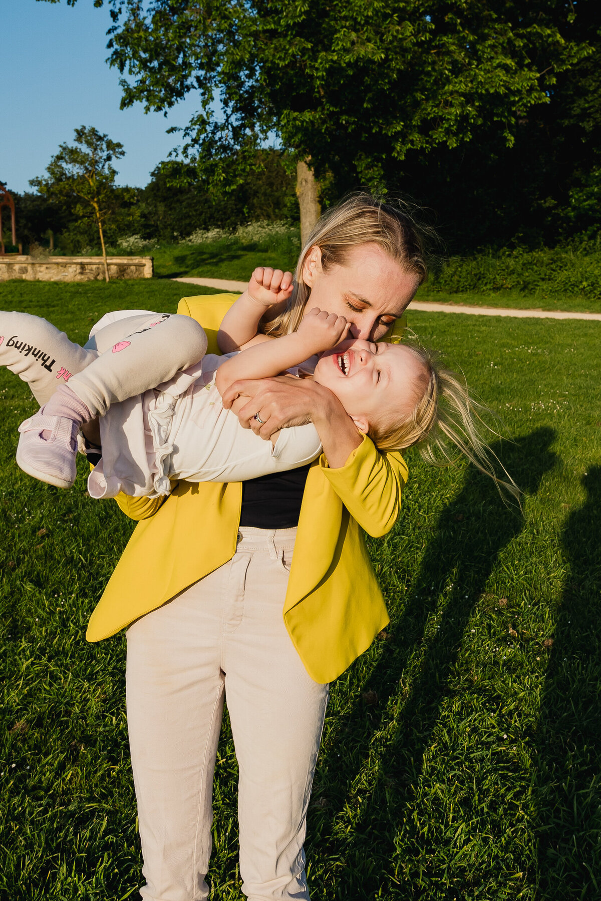 A woman in a yellow blazer lovingly lifting and kissing a laughing toddler in a park.