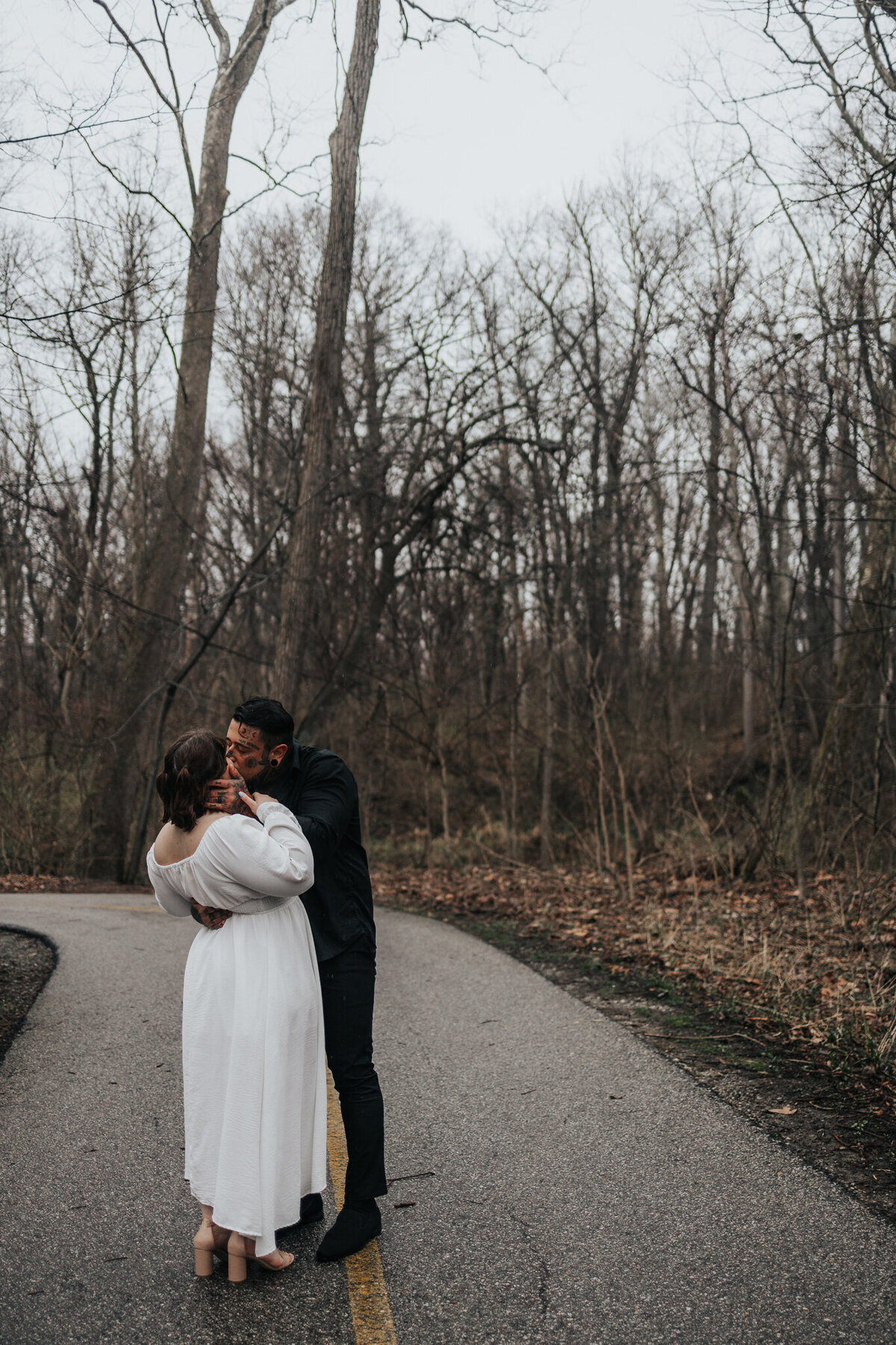 Couple shares romantic kiss during rainy engagement session in wooded Indiana park.