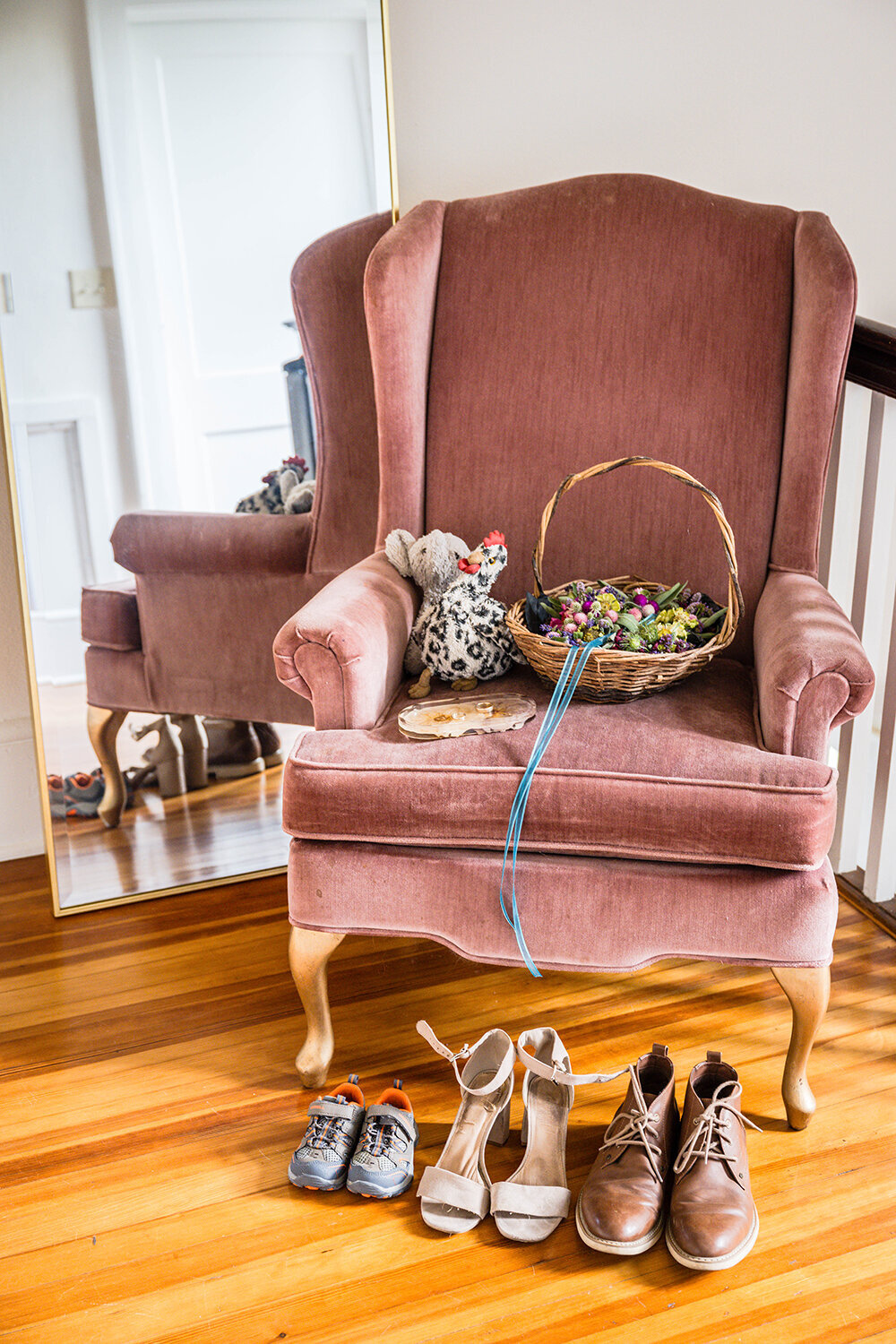 A pink antique chair sits in front of a mirror and holds a basket with three flower crowns, a ring dish with two rings, and two stuffed animals, a chicken and an elephant. In front of the chair on the wood floor are three pairs of shoes, growing in size as you look from left to right. The first pair is a pair of toddler hiking shoes, the second is a pair of high heels, and the third is a pair of brown dress shoes.
