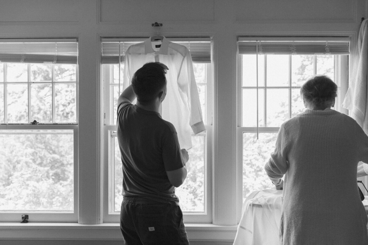 A groom prepares his wedding shirt with help from a loved one by the window—an intimate behind-the-scenes moment in Seattle wedding photography.