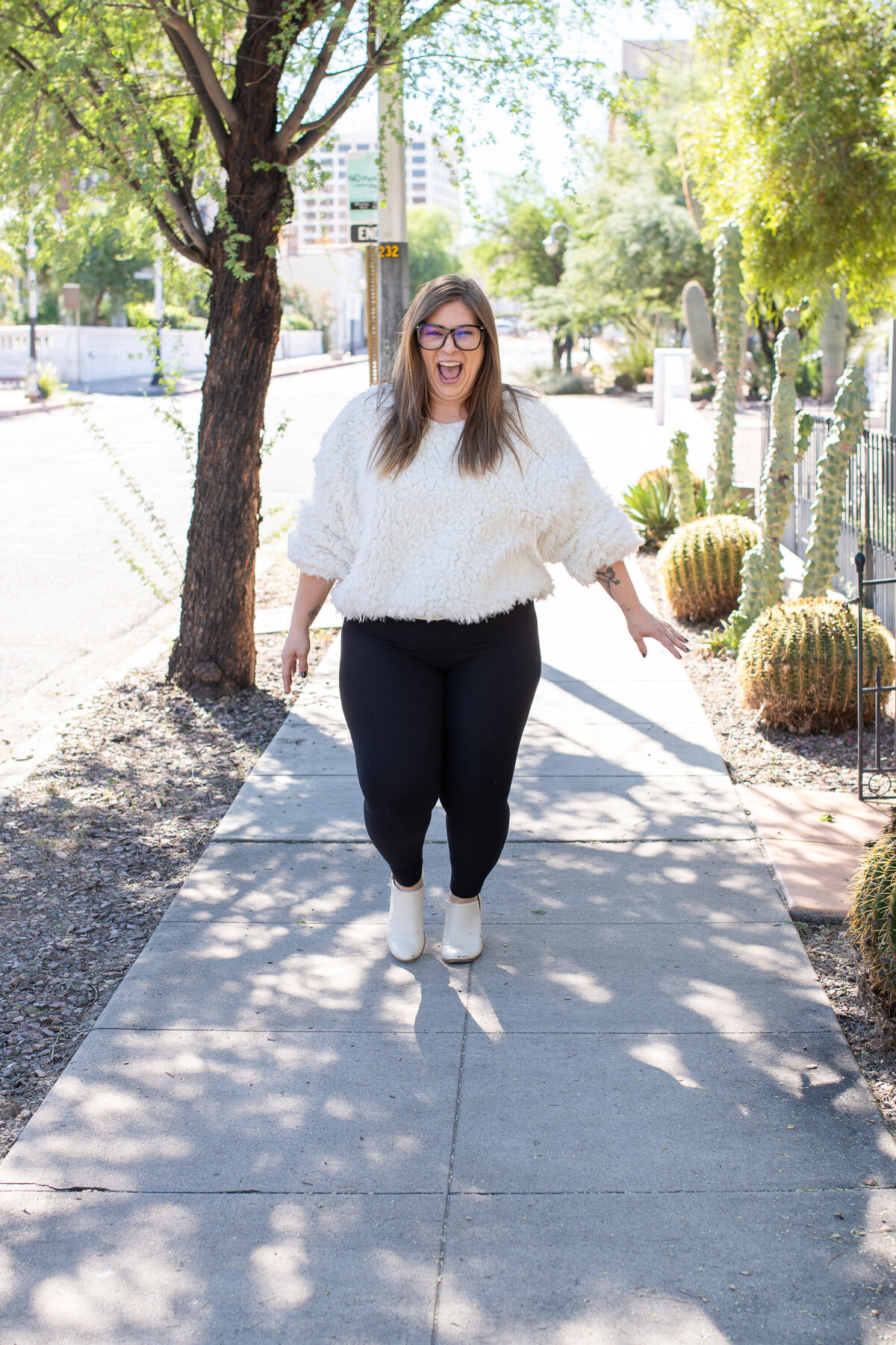Woman joyfully walking on a sunny sidewalk surrounded by desert plants and trees, photographed by Vyrl Photo, showcasing Tucson brand photography.