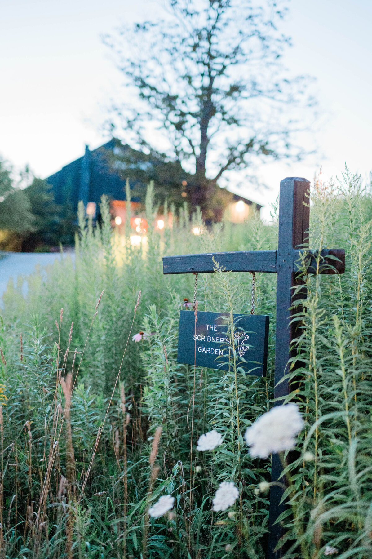 A wooden post holds a dark green sign reading “The Scribner’s Garden” amid tall wildflowers, with a warmly lit building in the distance.