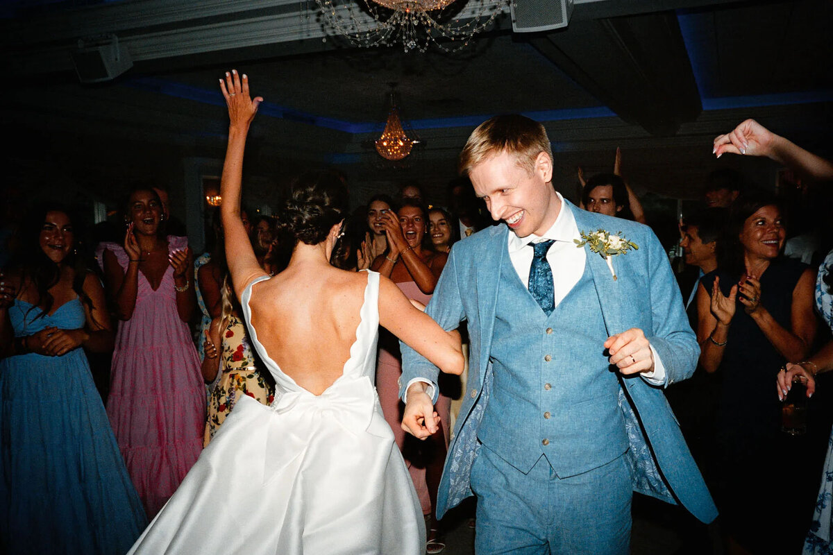 A bride in a white dress and a groom in a blue suit dance joyfully at their wedding, surrounded by cheering guests under chandeliers, as an NJ wedding photographer captures every magical moment.