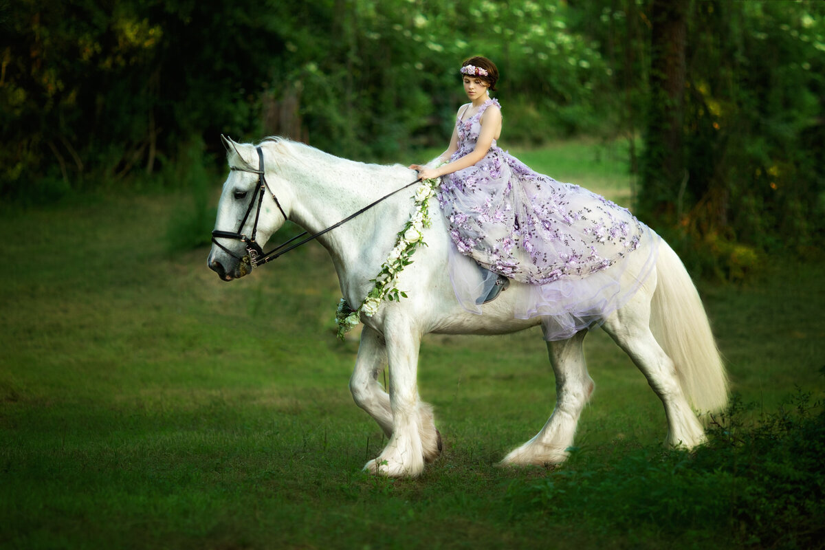 Female on a white Shire horse walking through a field.  The horse is wearing a floral garland from Fat Cat Flowers.  The girl is wearing Violet dress from Anna Triant Couture.  She has very short brown hair.  The background is green.