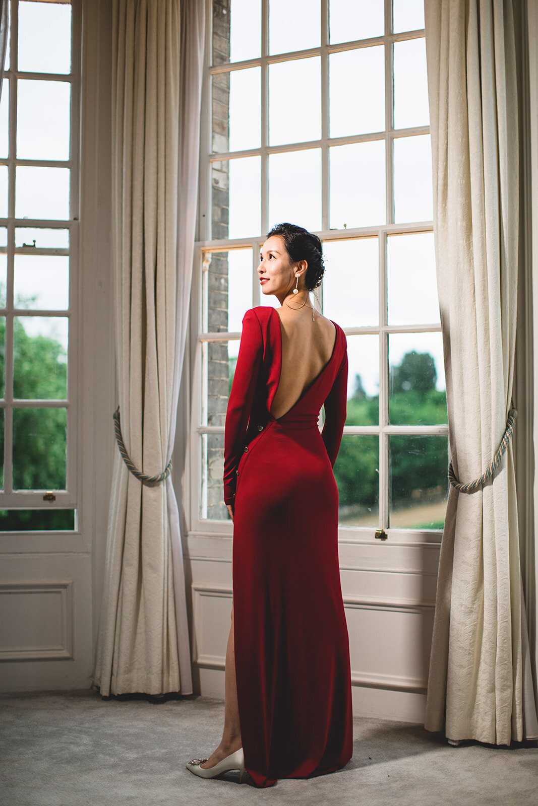 The Chinese bride dressed in red is lit and standing by a window at Claridges