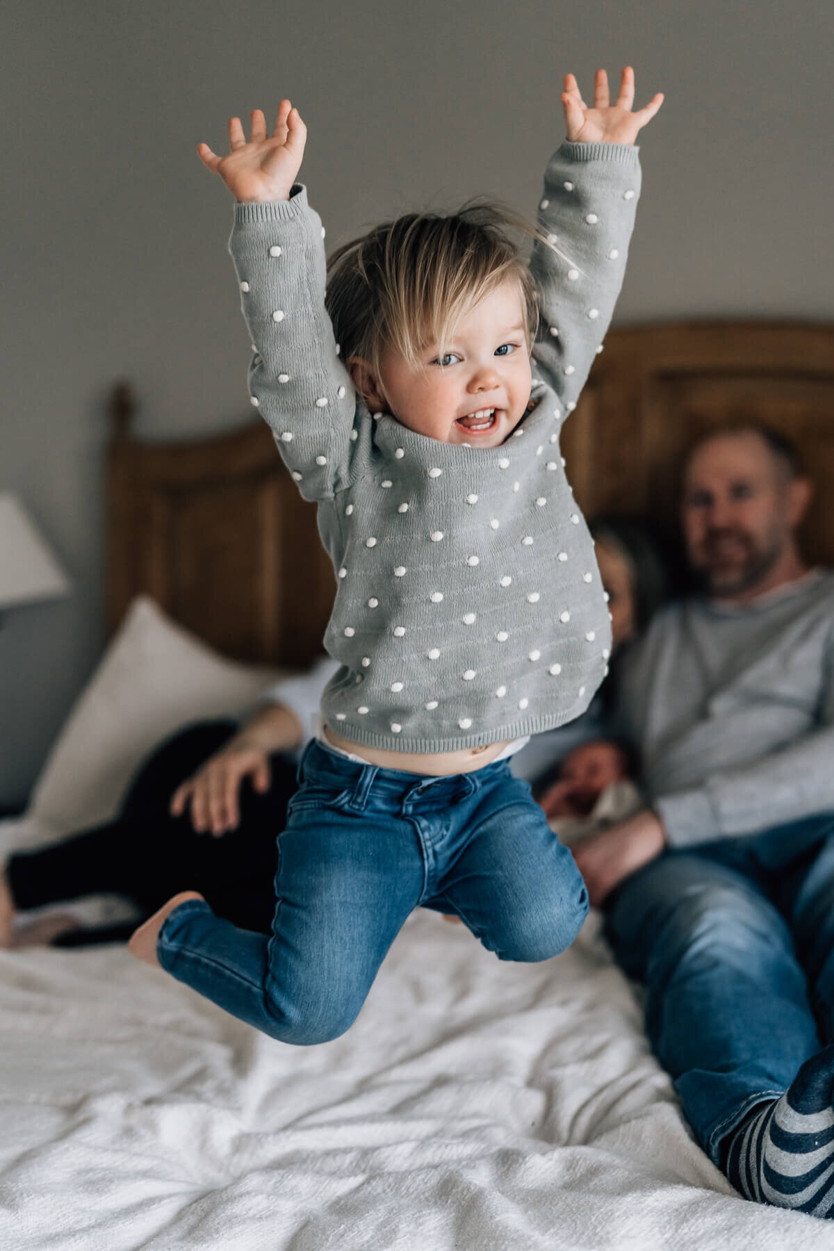 A two year old holds her hands above her head in mid air, while jumping on the bed during an in home newborn photography session.