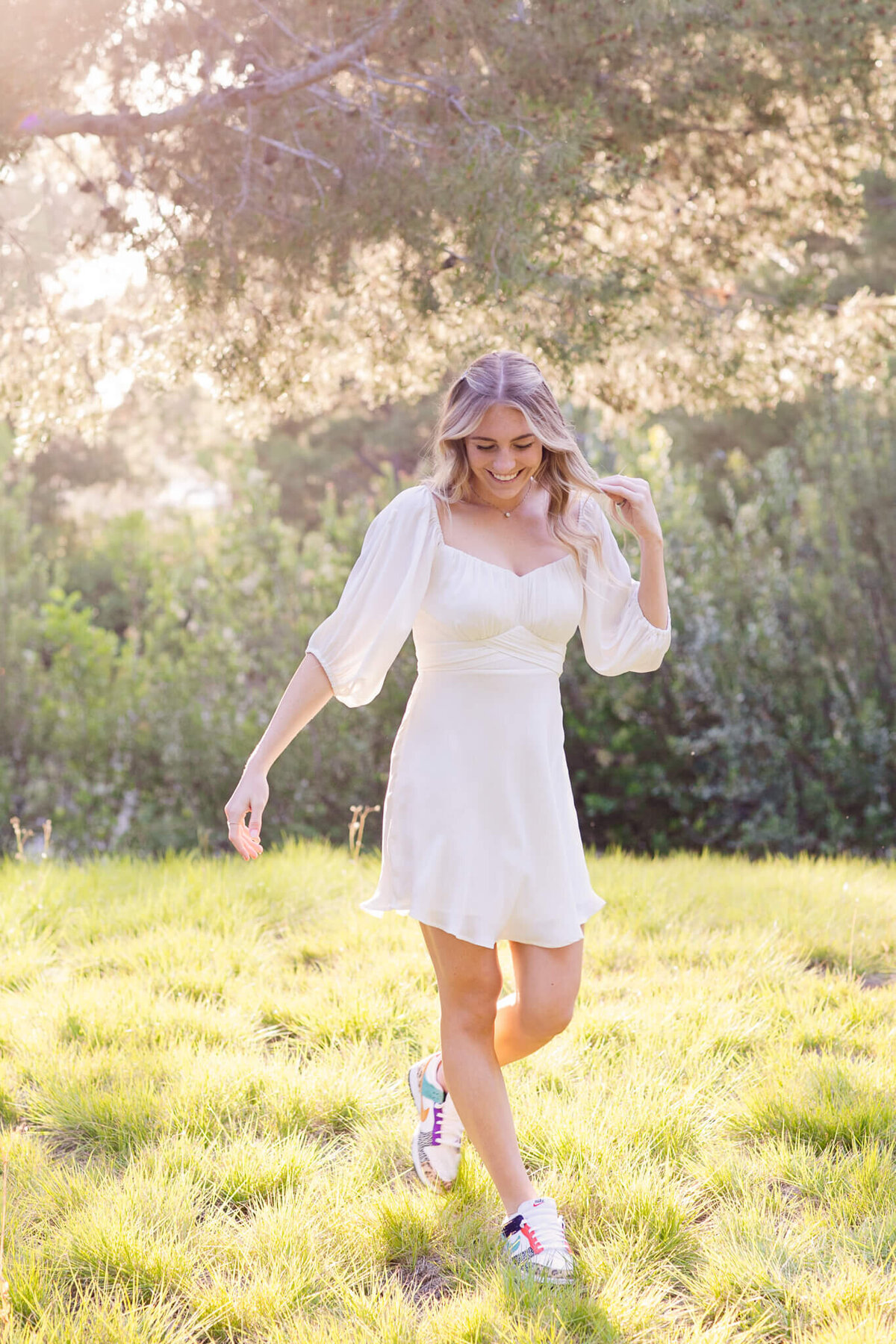 A woman in a white dress and sneakers walks joyfully on a sunlit grassy field, surrounded by green foliage. The sunlight creates a warm, serene atmosphere.