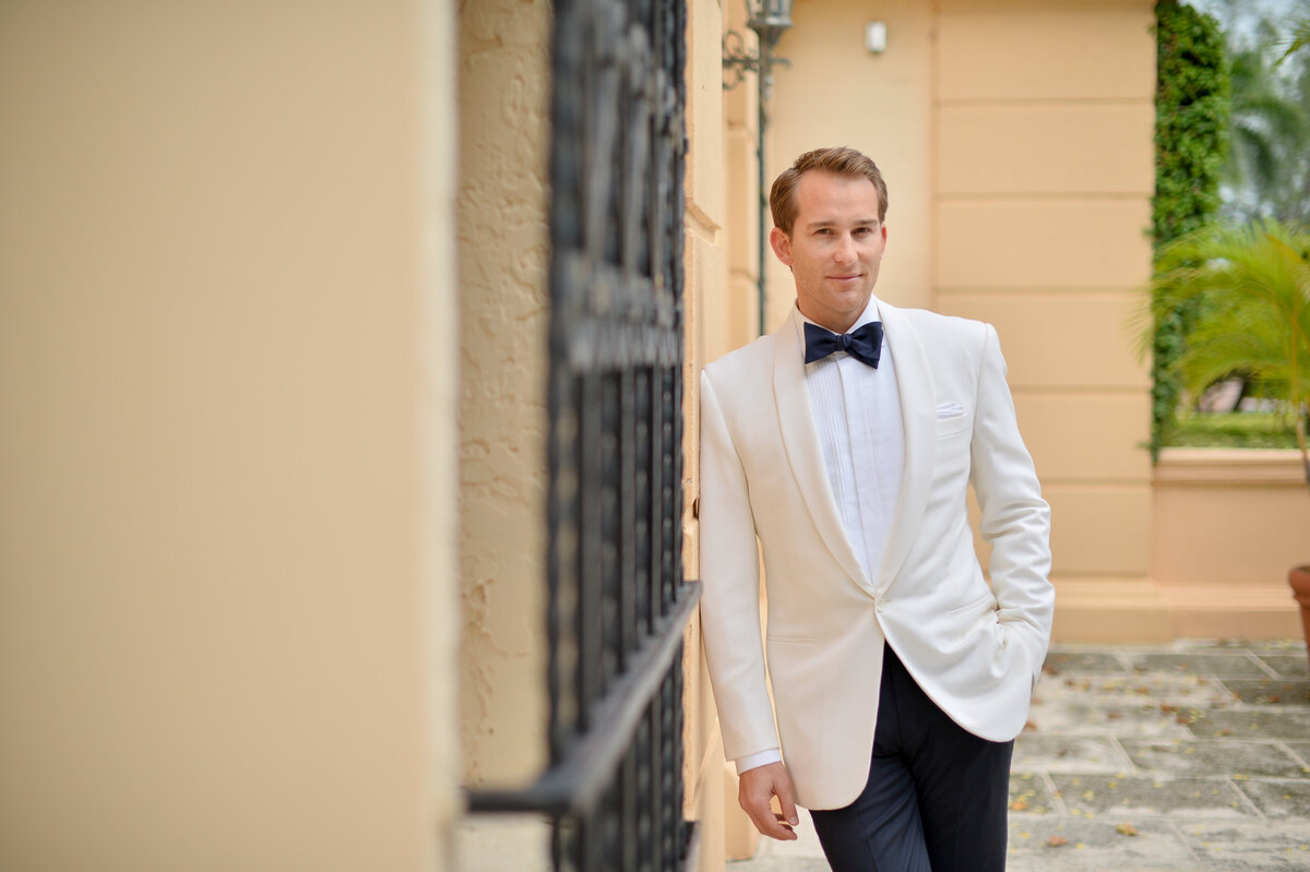 Groom in an elegant white tuxedo leaning against a wall, captured by Rodrigo Varela Photography.
