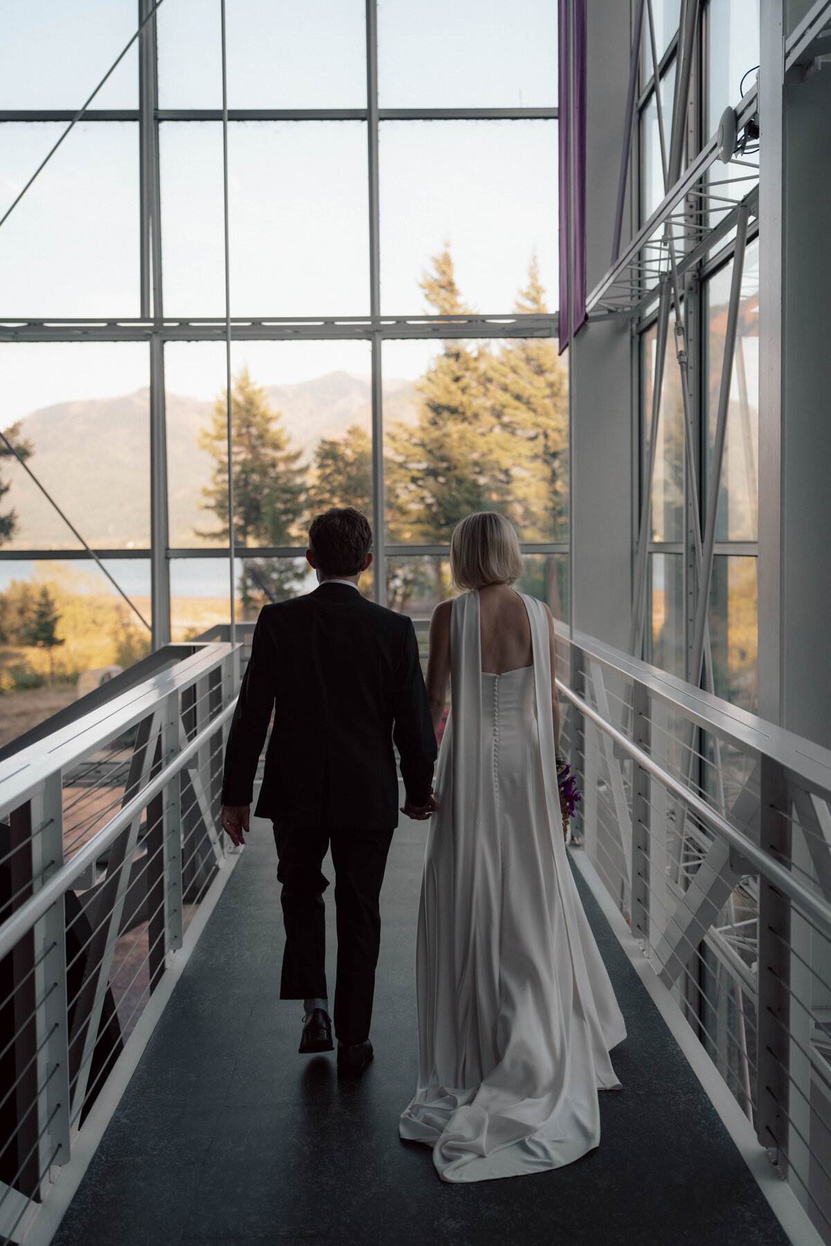 Couple holding hands walking through modern glass hallway with mountain view
