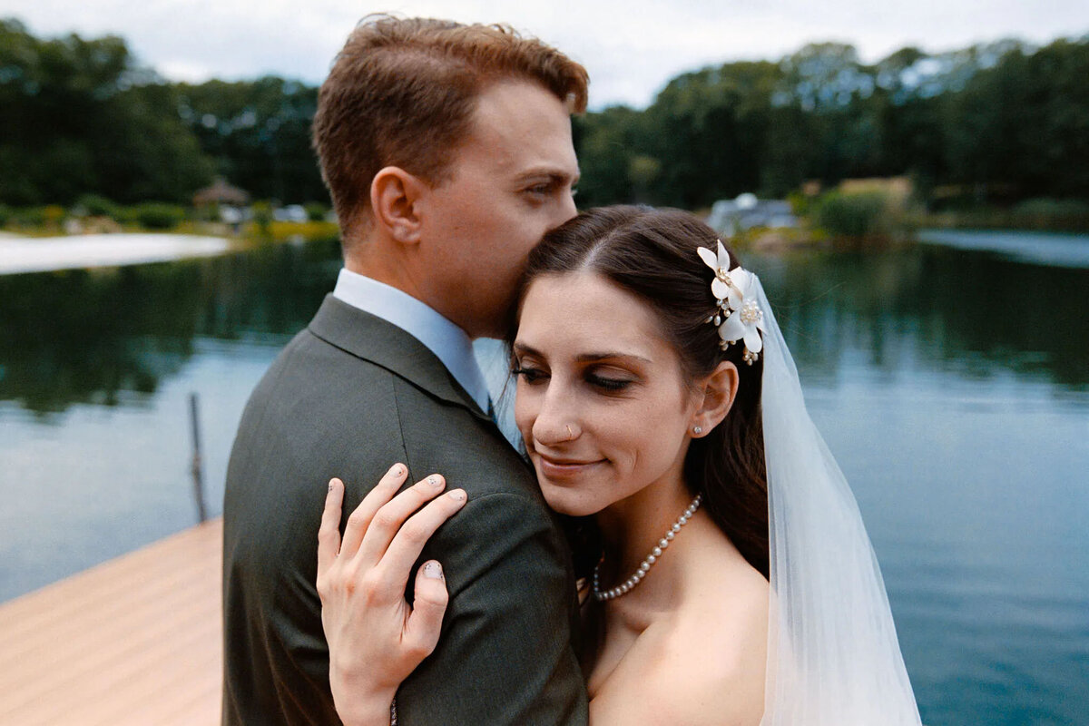 A bride and groom embrace on a dock by a calm lake. Captured by a film photographer NJ, the bride smiles softly with eyes closed, her veil and floral hairpiece glowing as she rests against the groom. Trees and water create a serene NJ wedding photographer backdrop.