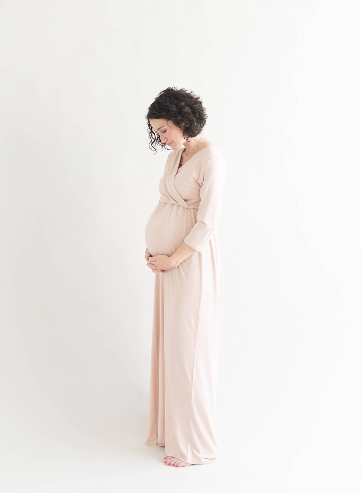 studio maternity portrait of beautiful mom in long cream dress on white background holding belly and looking down
