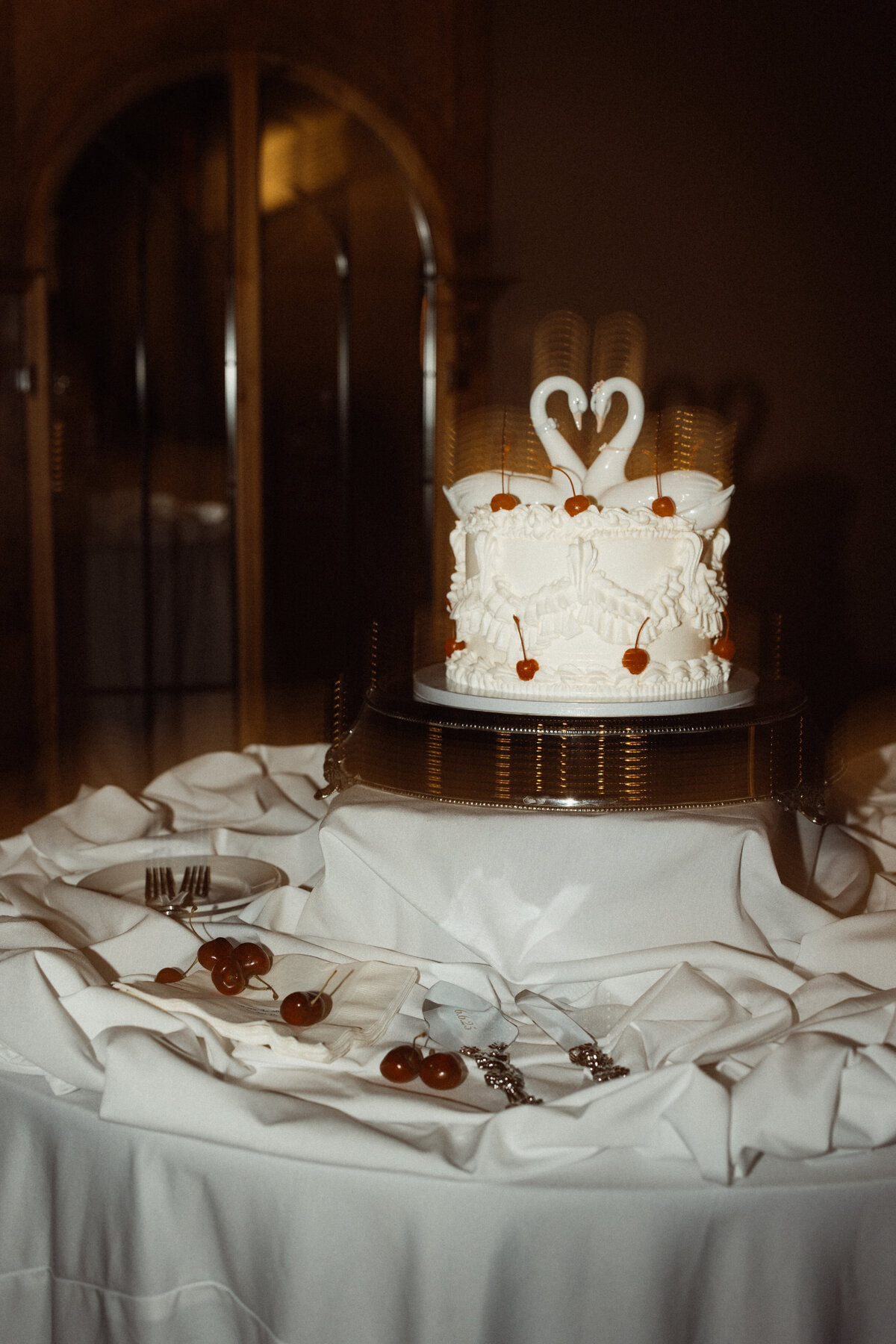 Vintage-inspired white wedding cake with swan toppers and maraschino cherry accents on a draped table at Le Venere, captured by Clewell Photography in Minnesota