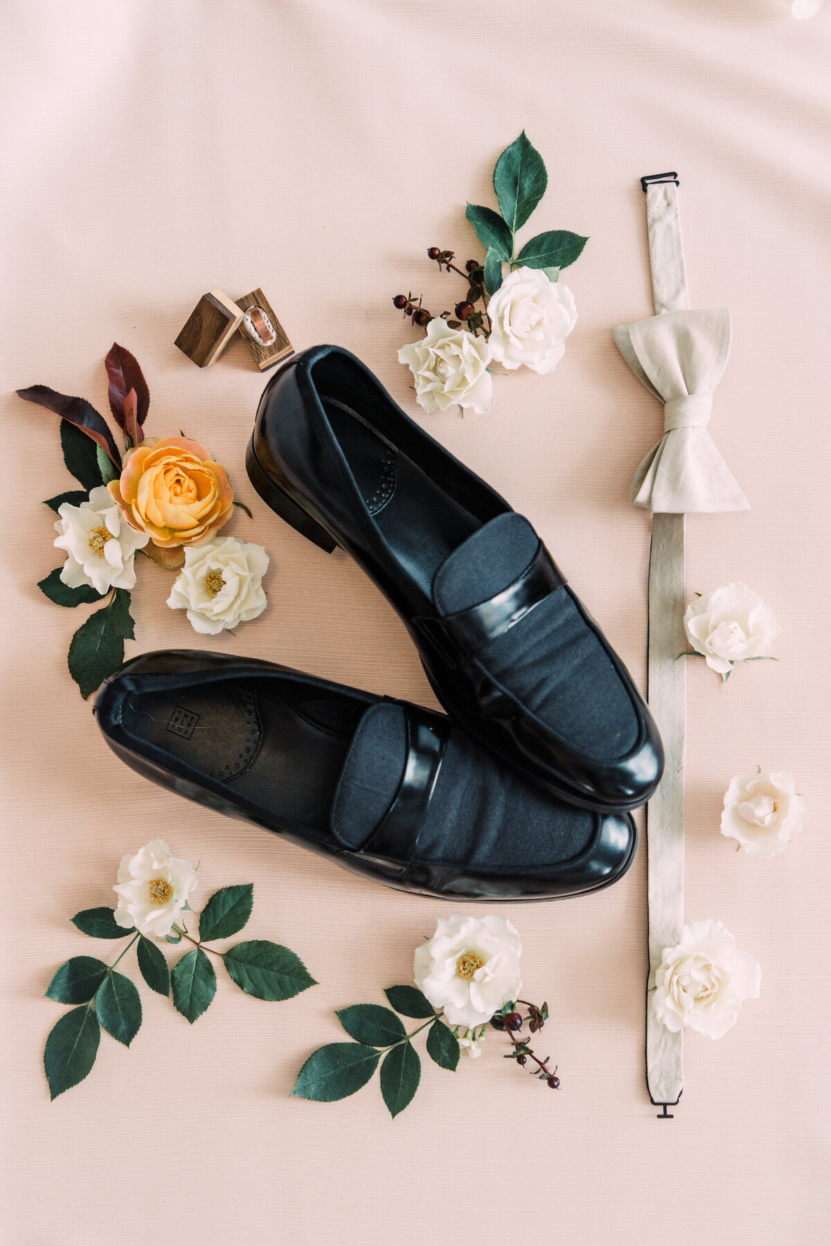 Styled wedding flatlay of groom's black loafers, white bow tie, and wood ring box with floral & greenery