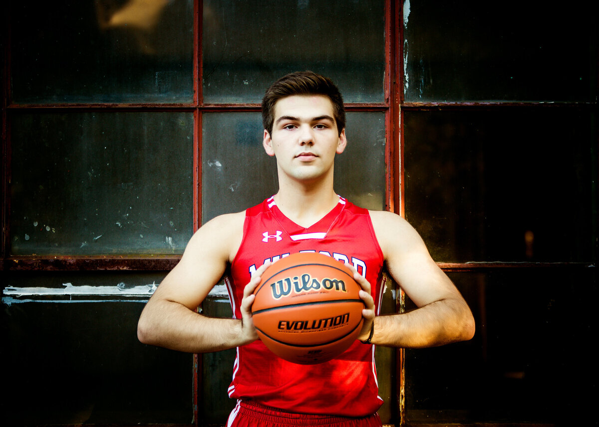 A male highschool senior wearing his basketball jersey holding a basketball in front of industrial window for a South Shore MA Portrait Photographer