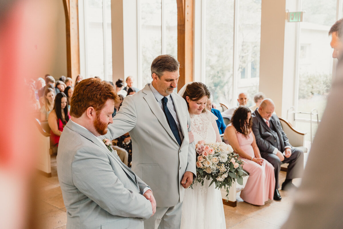 Emotional moment during a wedding at Ashton Gardens North, Houston, as the father of the bride gives away his daughter, holding hands in prayer. The groom stands beside them, and guests bow their heads in prayers  Captured by ShutterUp Photography, Houston wedding and event photographer.