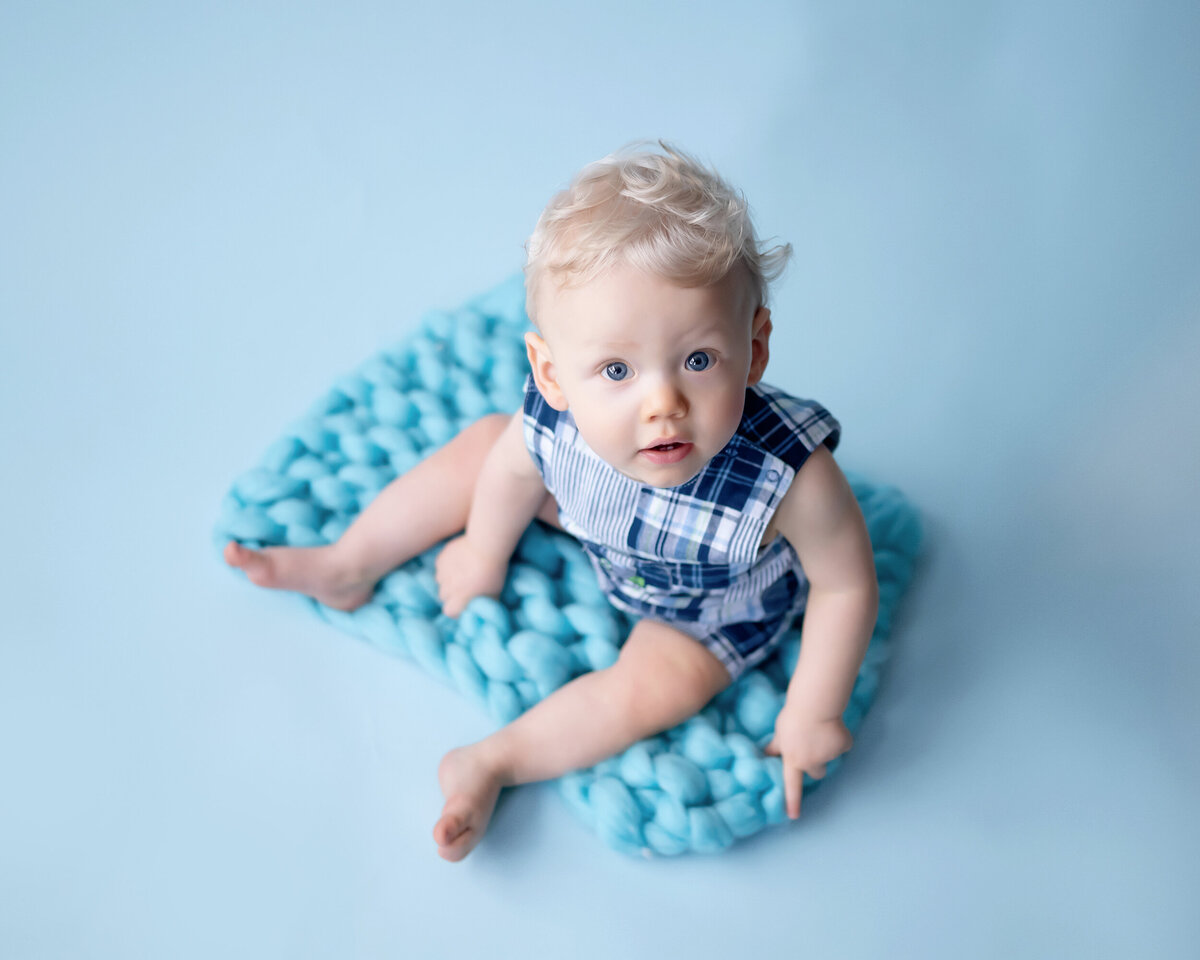 A baby in a plaid outfit sitting on a chunky blue knit blanket against a light blue background.