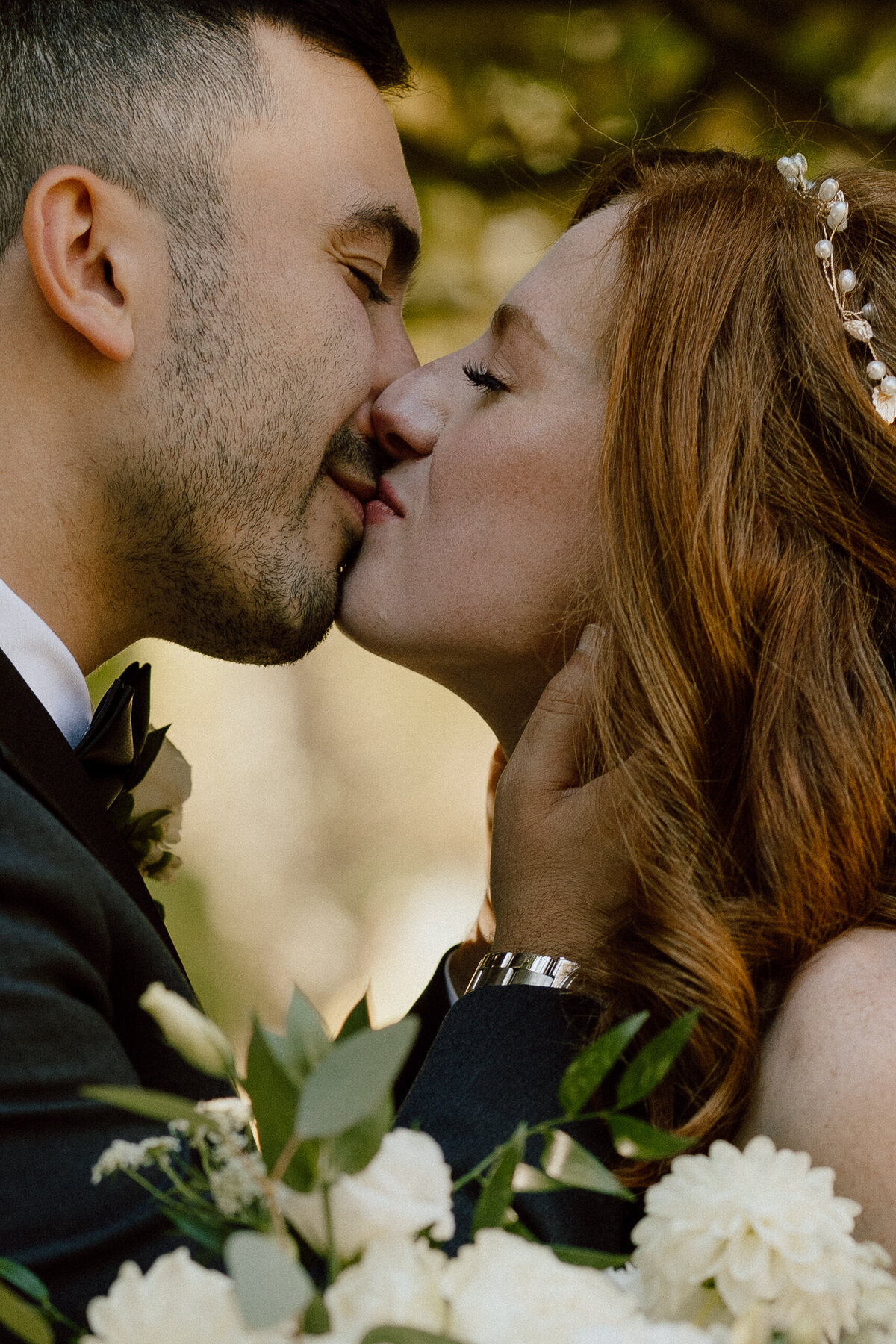 Close-up of a bride and groom sharing an intimate kiss on their wedding day. The bride, with red hair and a delicate hairpiece, and the groom, in a black tuxedo with a white boutonniere, are captured in a tender moment. The groom's hand gently cups the bride's face, and a bouquet of white flowers with green foliage is visible in the foreground, adding to the romantic atmosphere of the photograph.
