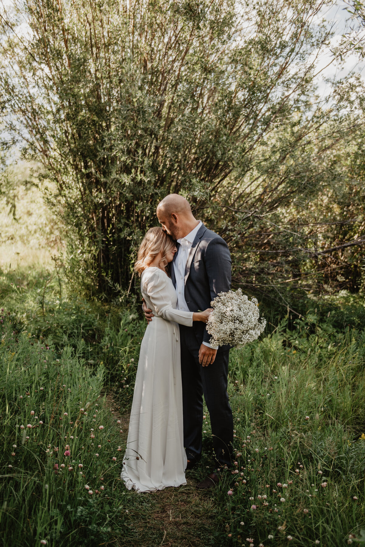 jackson wyoming photographer photographs bride and groom on their Jackson Hole wedding day holding each other and the groom is kissing the brides head as she holding her wedding bouquet