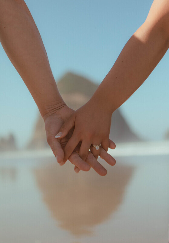 A couple holding hands with Haystack Rock blurred in the distance behind.