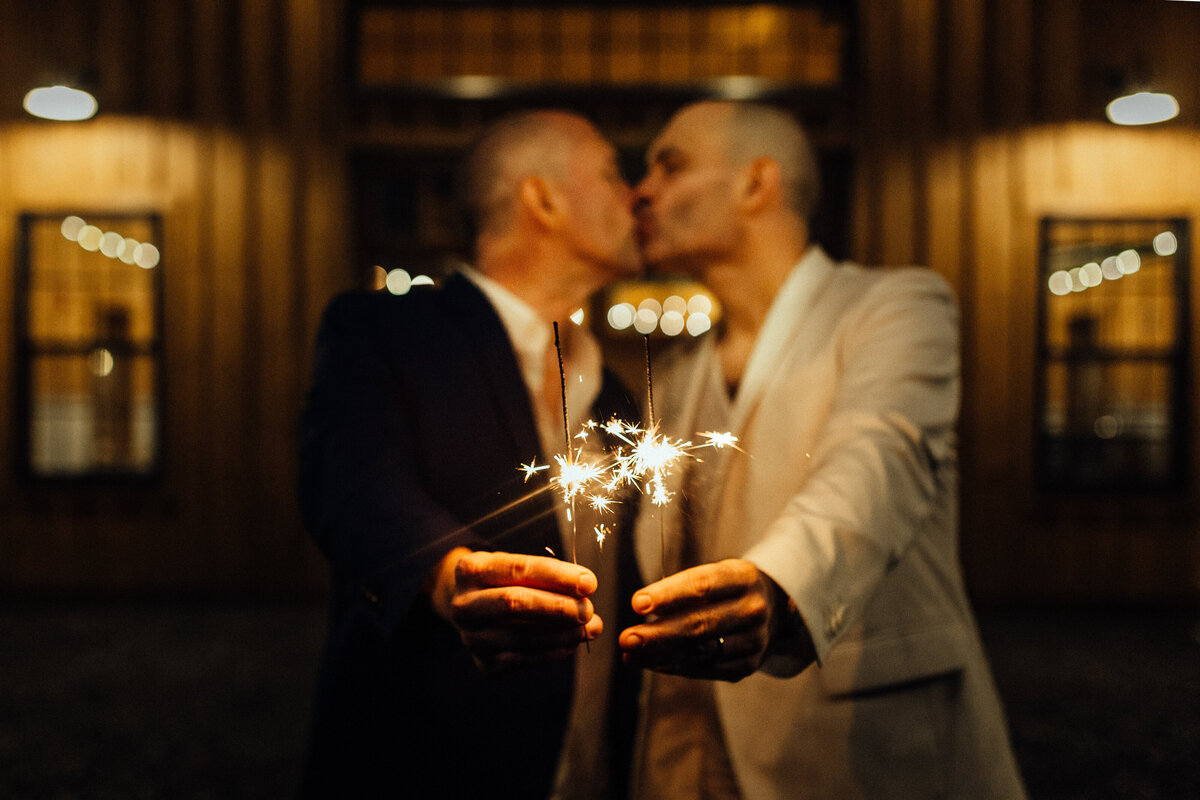 Two grooms kissing while holding out sparklers.