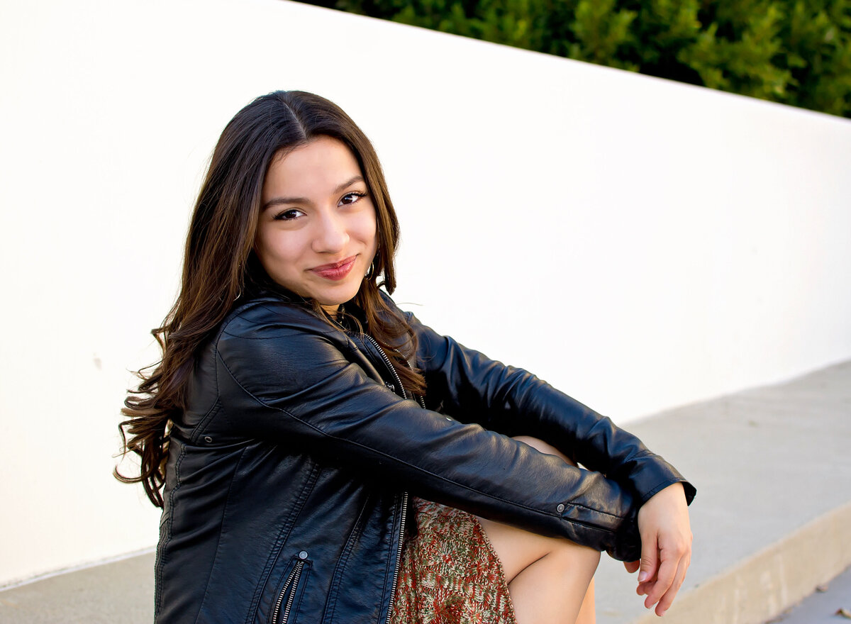 Senior girl sitting on the curb in a black leather jacket smiling at the camera
